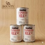 12 stacked cans of Kirkland Signature Evaporated Milk (354mL) with Vitamin D on a clean, light wood background for The Bounty Shop.